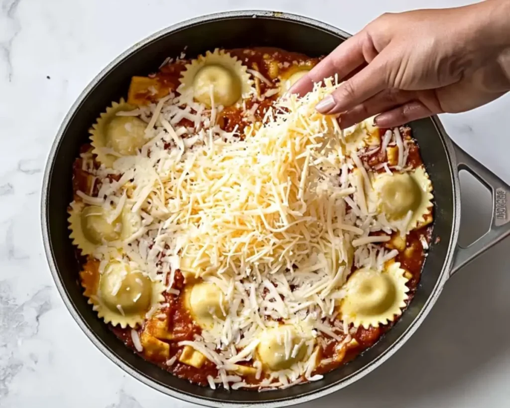 A hand sprinkling shredded cheese over cheese ravioli in a skillet.