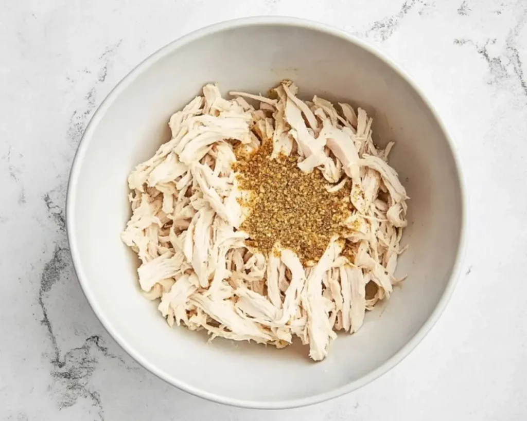 Shredded chicken tossed with aromatic spices in a mixing bowl
