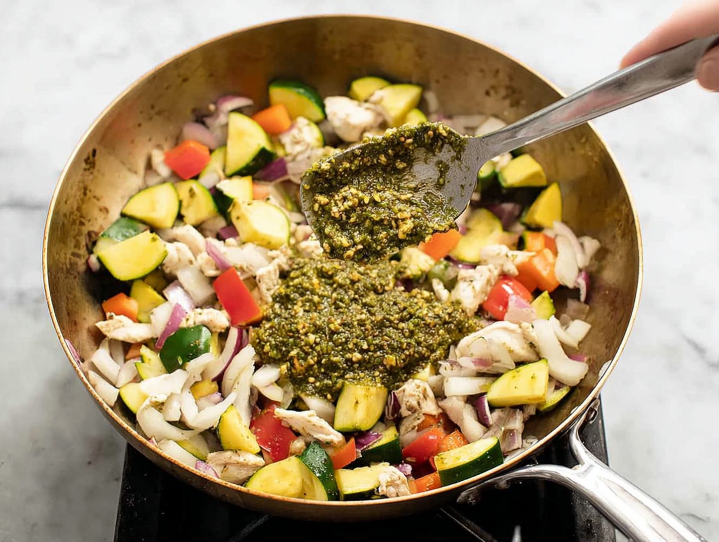 Pesto sauce being mixed into skillet with chicken and vegetables